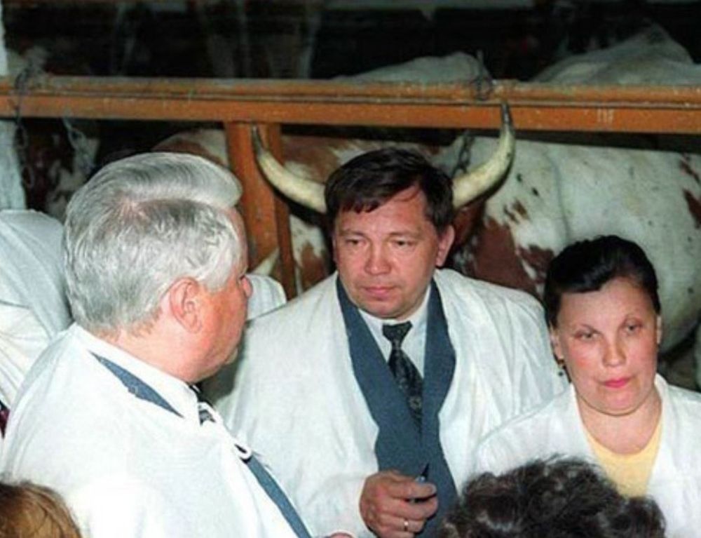 The image shows a group of people standing together inside what appears to be a barn or livestock enclosure. They are wearing white coats, suggesting a professional or scientific setting, possibly veterinarians or animal experts. In the background, there is a cow with large, curved horns standing behind the people. Due to the positioning of the cow, the horns are aligned with the head of the person in the center, creating the humorous visual illusion that the person has horns.