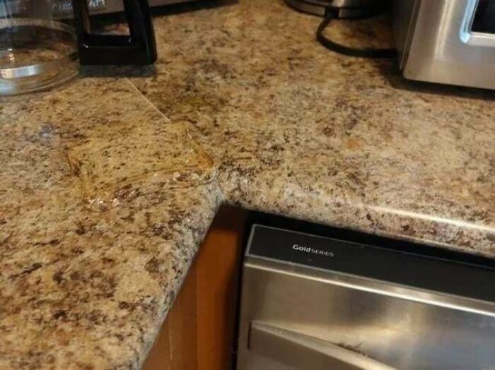 This image shows a kitchen countertop made of mottled brown and beige material. On the counter, in the left corner, there is a piece of bread with jelly or jam on it. The bread blends in almost perfectly with the color and texture of the countertop, making it very hard to see. Only the glossy sheen of the jelly helps the bread stand out from the background. This image demonstrates how similar colors and textures can create visual camouflage, making objects difficult to spot.