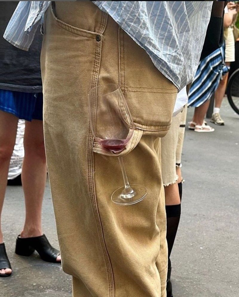 The image shows a close-up of a person's hip and upper leg area. The person is wearing light brown pants with a slanted side pocket. Unusually, a wine glass with a small amount of red wine is resting perfectly in the pocket, standing upright as if the pocket was designed to hold the glass. The background is out of focus, showing other people's legs and feet, indicating an outdoor social gathering. The scene is humorous as pants pockets are not typically used (or shaped) for holding delicate objects like wine glasses.