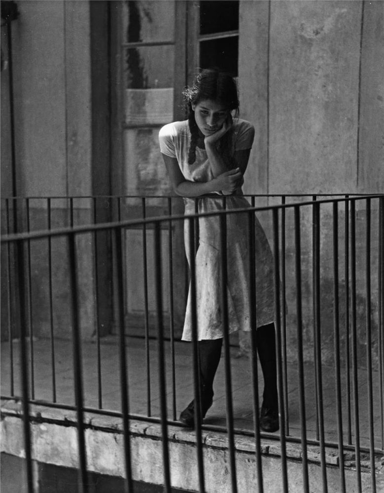 A b&w image  of a young woman leaning on a balcony gazing wistfully below. 