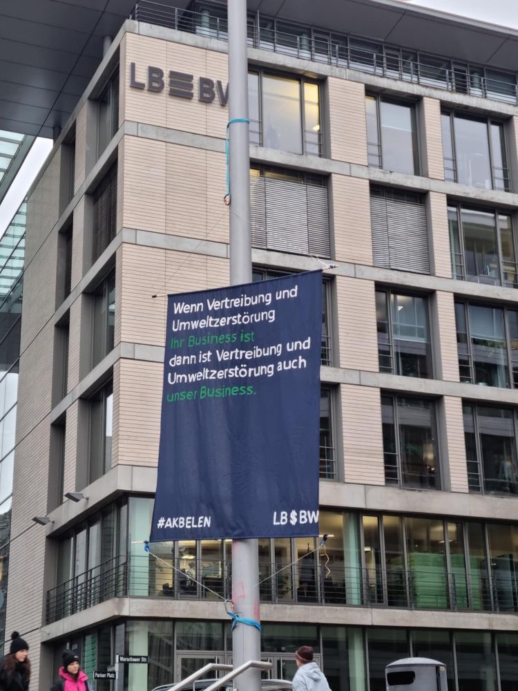 Banner an einem Laternenmast im Stil einer LBBW Werbung. Im Hintergrund die LBBW. Auf dem Banner steht "Wenn Vertreibung und Umweltzerstörung ihr Business ist dann ist Vertreibung und Umweltzerstörung auch unser Business. Das LBBW Logo ist verändert und enthält ein $