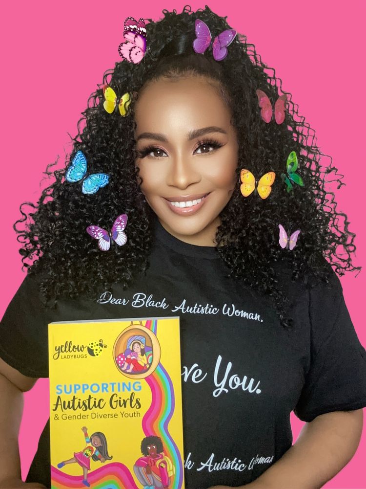 Phenomenally autistic holding a copy of supporting autistic girls book the cover is yellow she is wearing a black T shirt a curly hairstyle with rainbow butterfly clips 