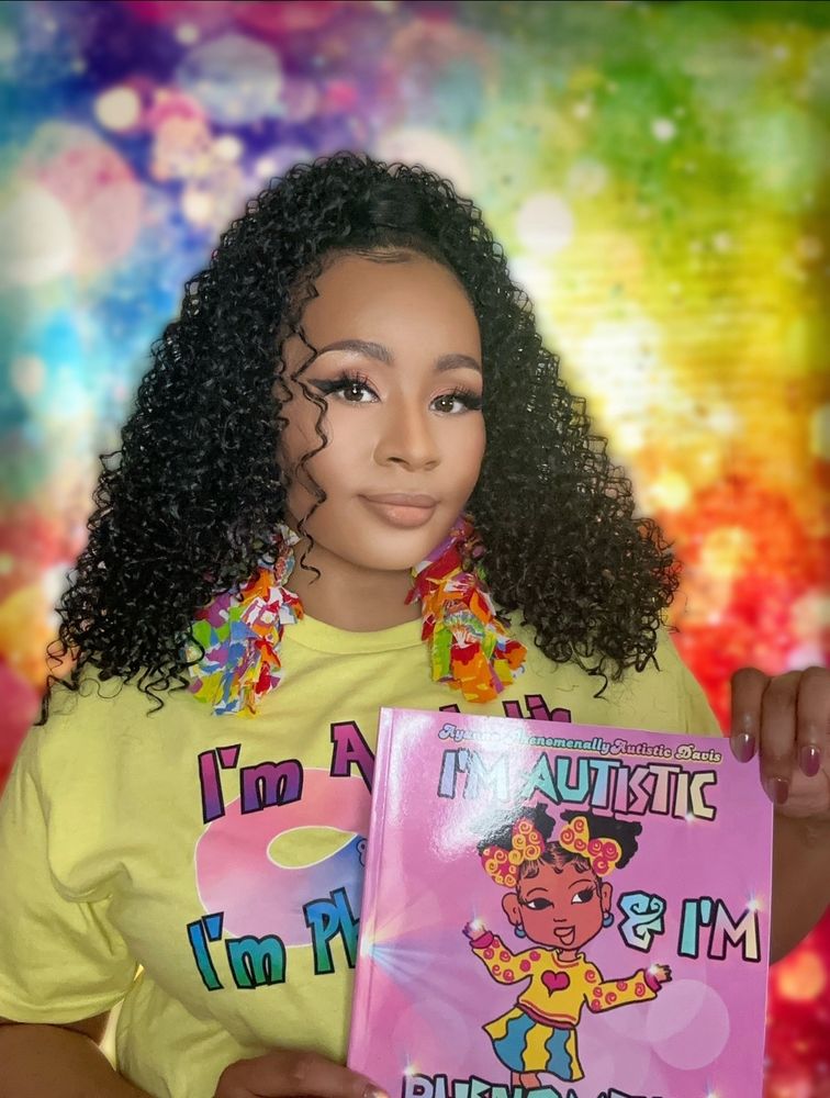 Phenomenally autistic holding her children’s book standing in front of a rainbow background she has on a yellow shirt 