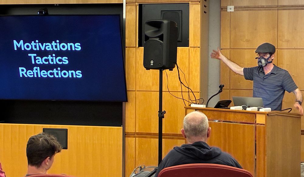Mark Dixon speaking at a podium wearing a GVS Elipse mask. Screen says "Motivations Tactics Reflections"