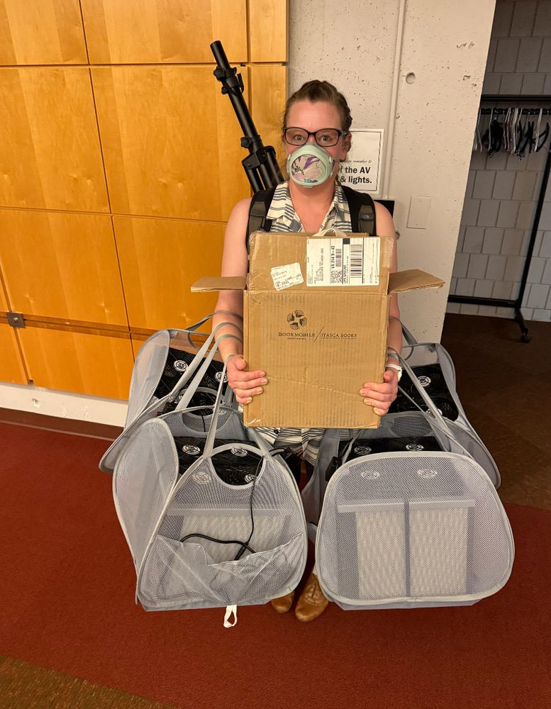 White female wearing Flo mask holding a box and four air filters in mesh bags. A speaker stand is in her backpack. 