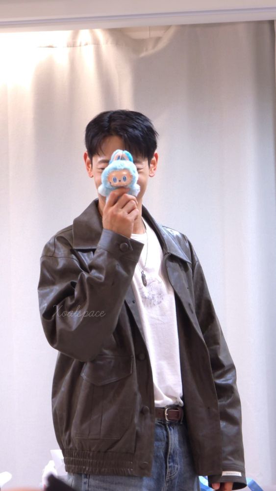 Minho from SHINee, dressed in a jacket and jeans, holding a labubu plush toy in front of his face. 