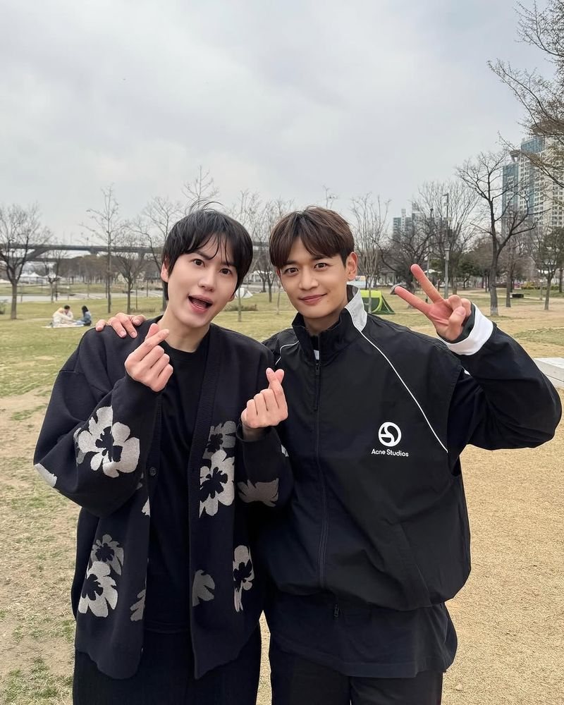 In a park, Minho from SHINee and Kyuhyun from SuperJunior stand side by side, smiling for the camera.