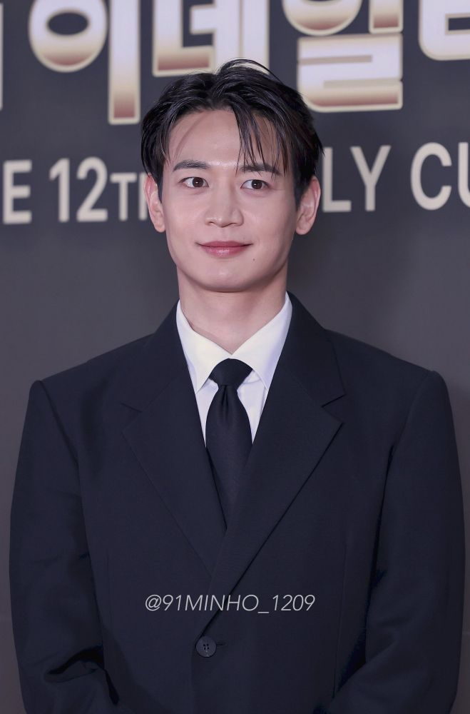 Minho from SHINee, dressed in a suit and tie, poses in front of a stylish wall backdrop.