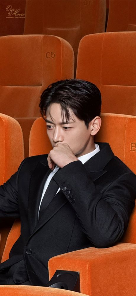 Minho from SHINee, dressed in a suit, relaxes in an orange chair, exuding a confident and friendly vibe.