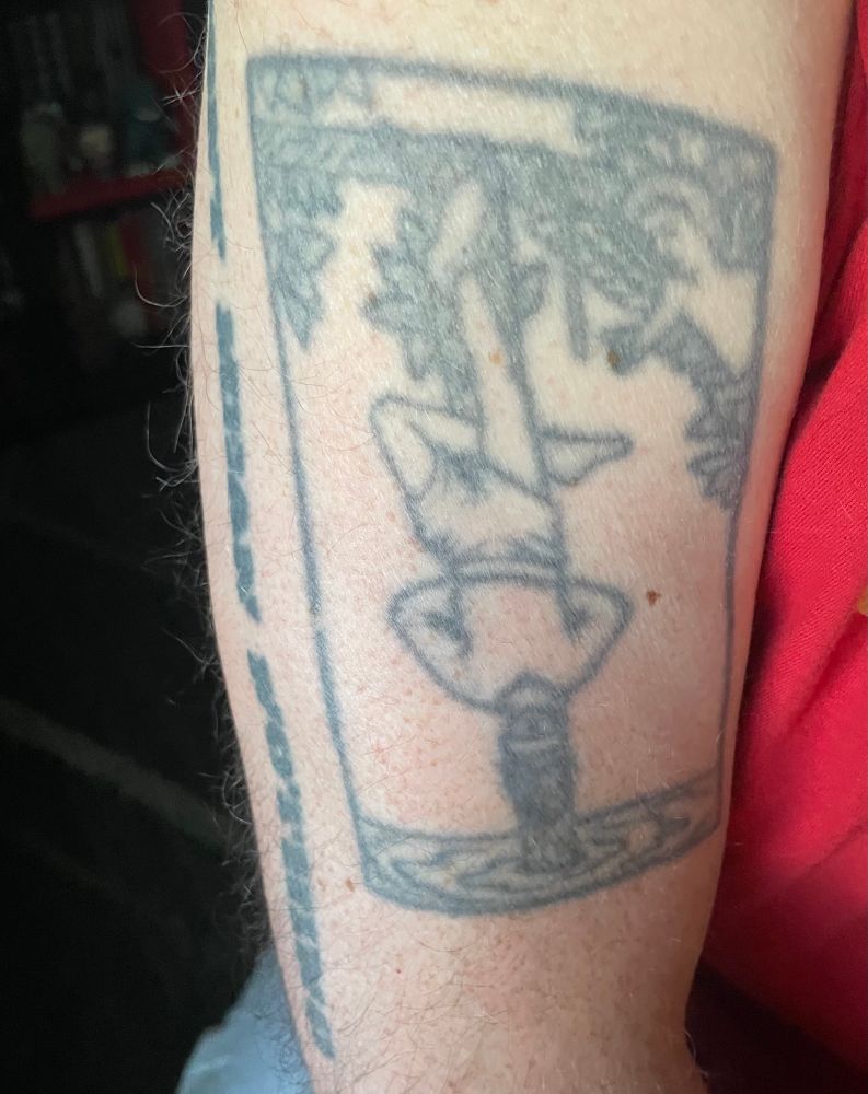 Washed-out Hangman tarot card tattoo on what looks like the leg of a small child but is in fact the arm of an adult man 
