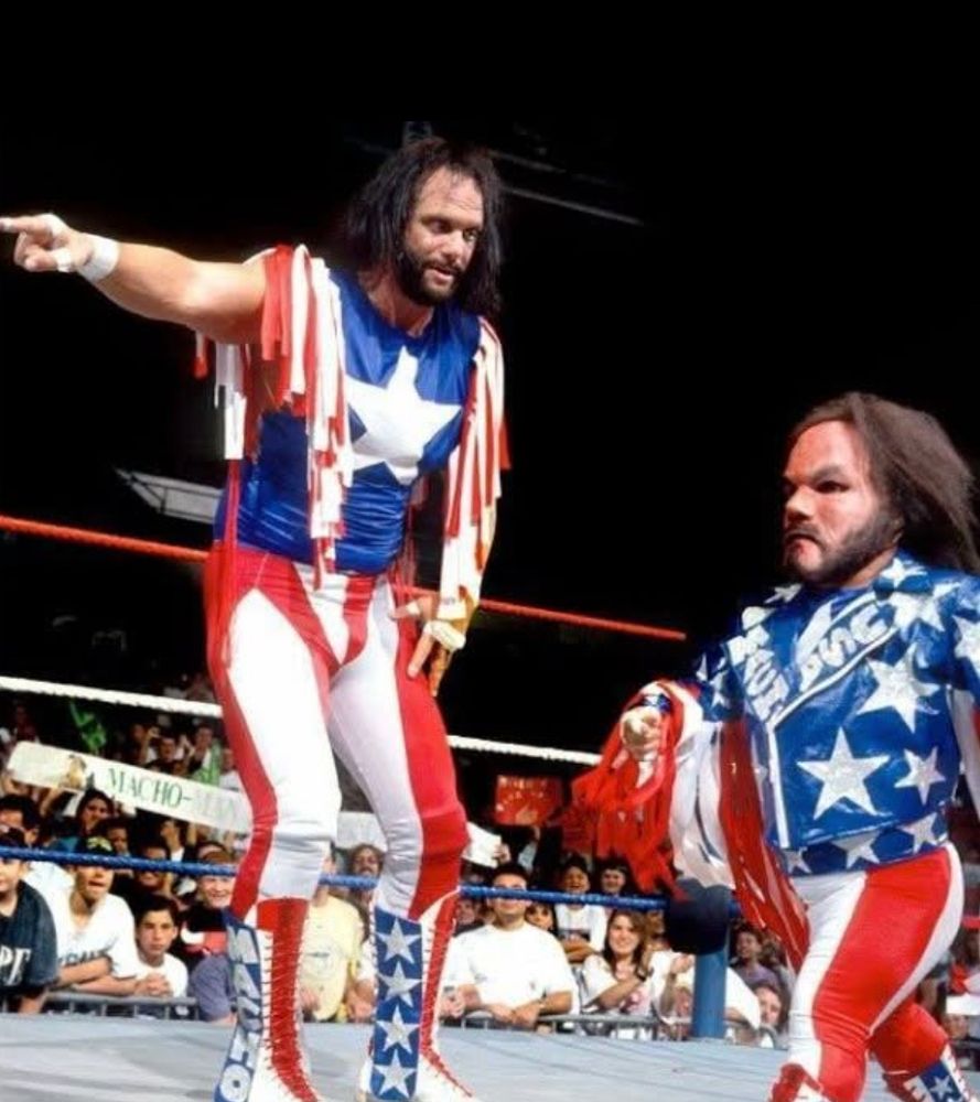 Macho Man Randy Savage, in a wrestling ring decked out in red white and blue Stars and Stripes gear, points forward directing a little person version of himself to step forward and get it done. 
