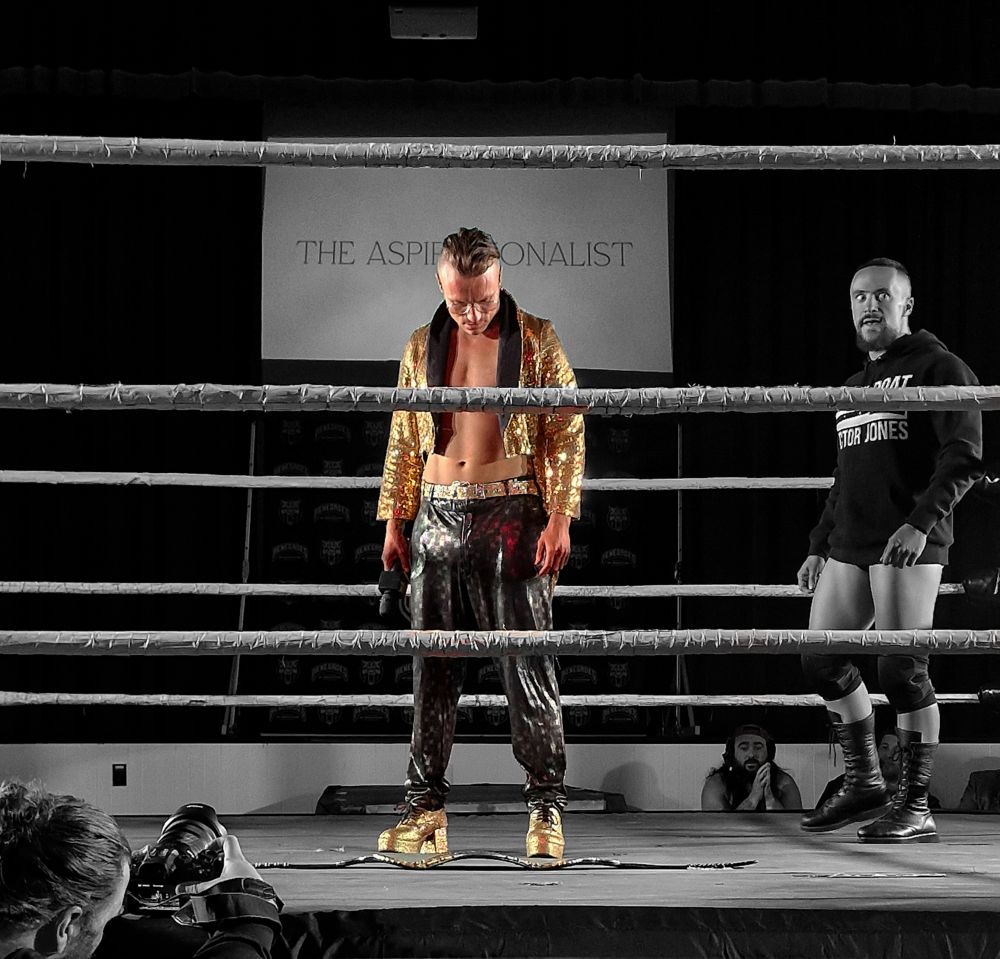 Lochy Hendricks, in a gold glittery jacket, looking down at the Renegades World Title lying in the middle of the ring
