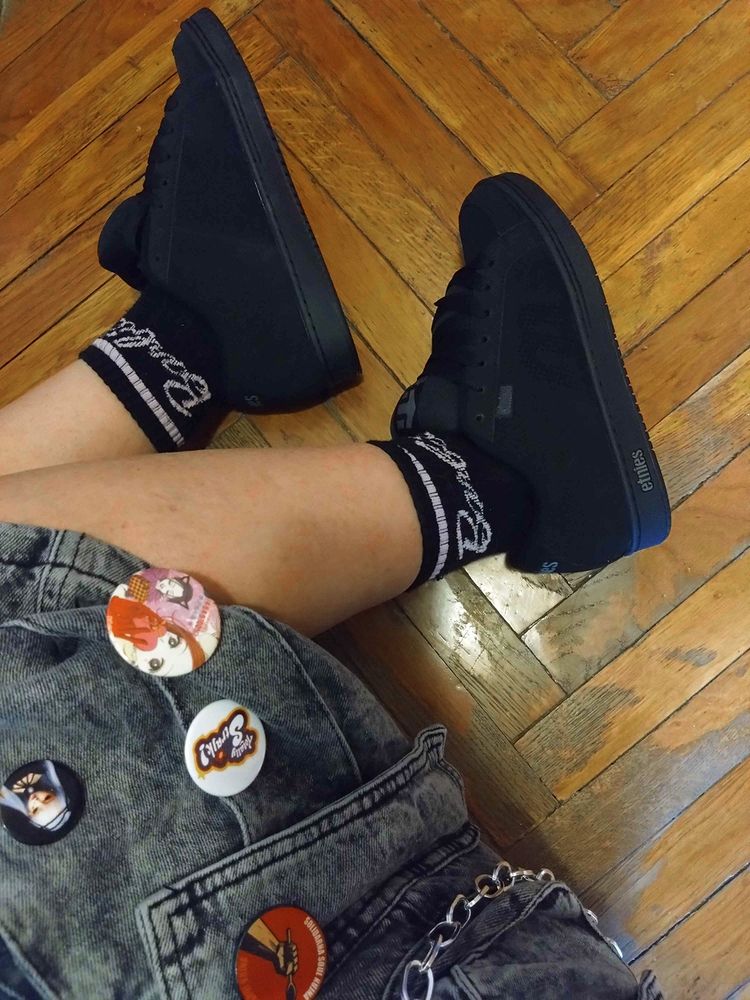 close up of black skate shoes, while sitting on floor. part of the cargo shorts is also visible with various pins on the big side pocket