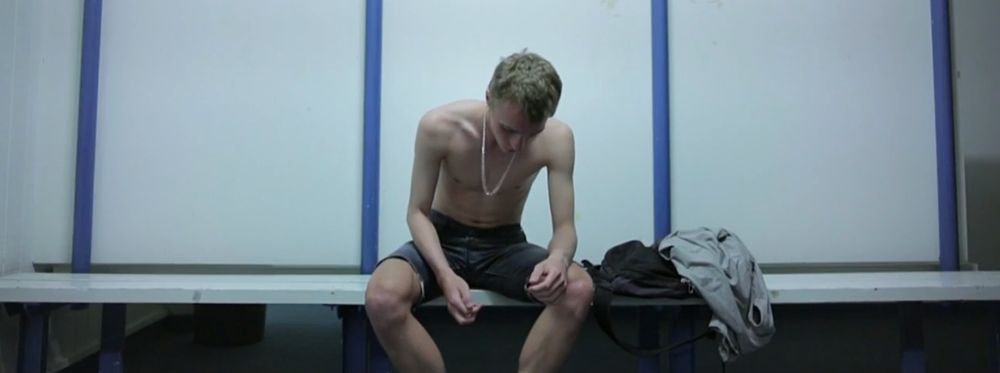 From the video for Lorde's Royals: a young man sits on a plain bench wearing shorts with some tops beside him, reaching down to untie his shoelaces. Behind him is a white wall with blue vertical stripes.
