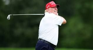 Donald Trump wearing a white polo shirt, red MAGA hat, watches his golf shot. He is holding a golf club in the follow-through position with the club head behind him. Taken at his Jupiter, FL golf course on April 5, 2025.