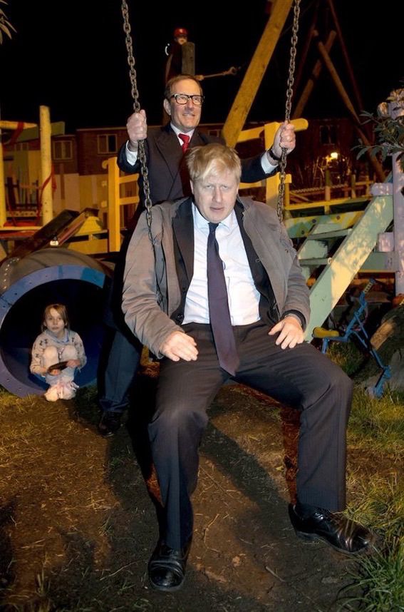 In a nightmarish scene, Boris Johnson in a dark children’s playground manspreads on a swing that is being pushed by a maniac in a double-breasted suit. Behind them, a small child huddles in the mouth of a tunnel contemplating future PTSD