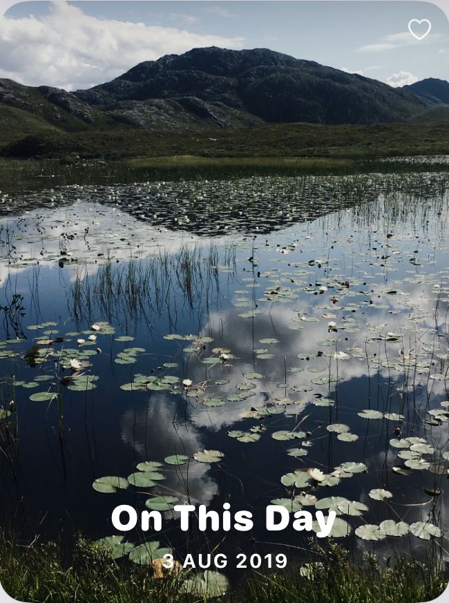 A mountain and clouds reflected in a loch full of water lillies. Automatically labelled “On This Day 3 August 2019”