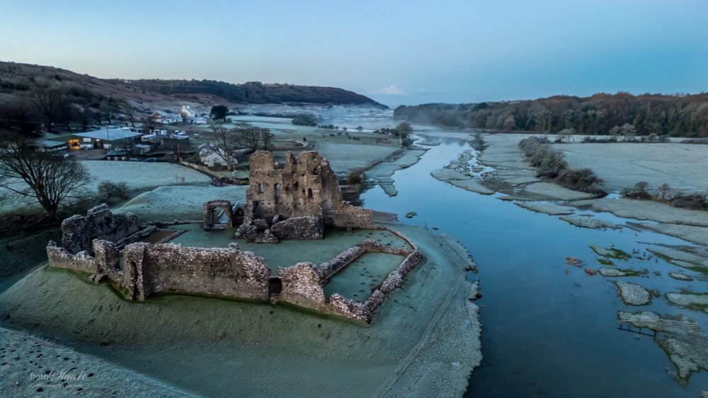 A frosty landscape featuring a historic stone ruin surrounded by a shallow moat or river, reflecting the cold morning light. The ruins, of Ogmore Castle castle, have crumbling walls and arches, sitting on a grass-covered island. In the background is a farm and small buildings nestled near the riverbank, with frosted fields stretching into the distance. Mist rises over the water, and the scene is framed by rolling hills and a line of trees under a pale blue sky.