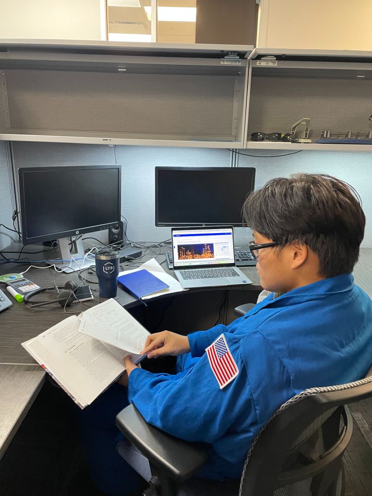 Bobby He wearing his LyondellBasell jump suit working at his desk while reading a book. 