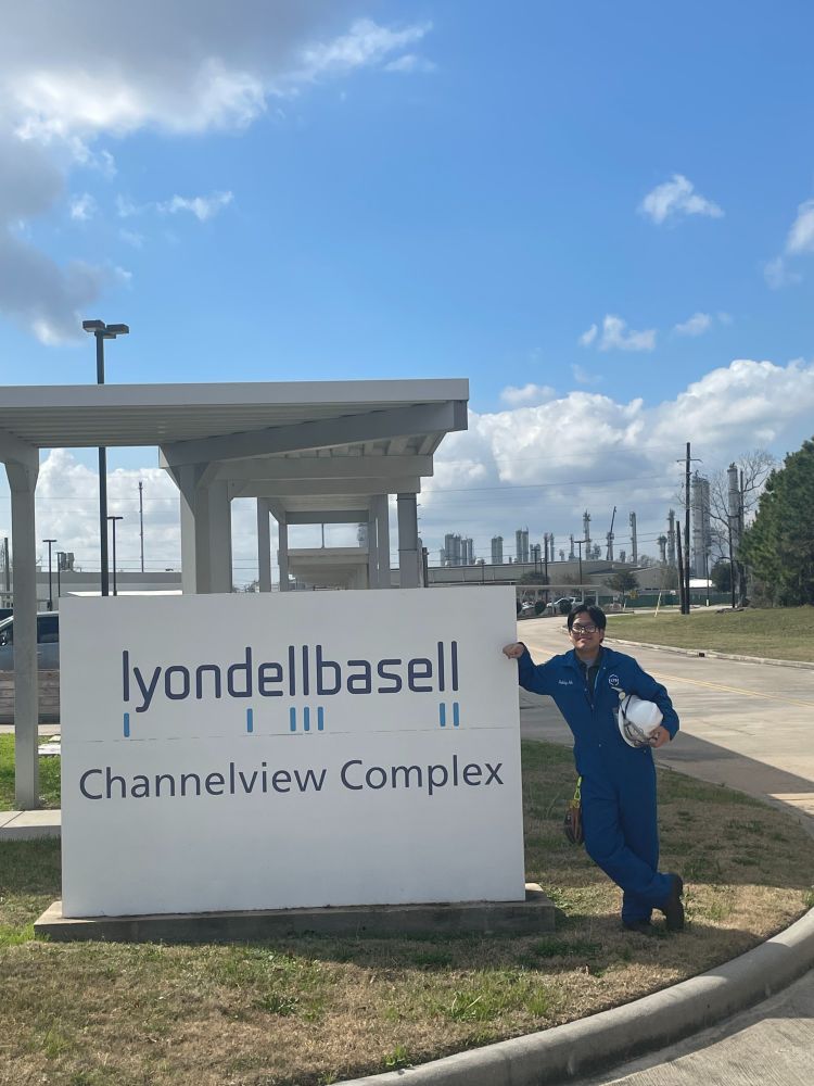 Bobby He leaning against the LyondellBasell Channelview Complex sign in his work wear jump suit. 