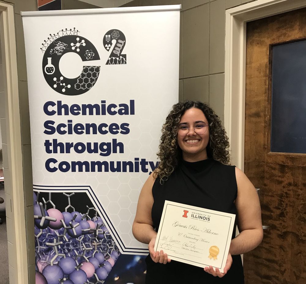 Génesis Ríos-Adorno poses with her award certificate in front of a C2 Chemical Sciences through community sign.
