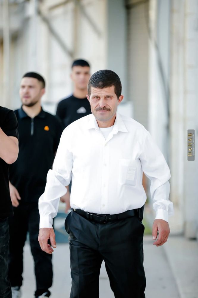 A man wearing a white button-down shirt and dark trousers walks forward with a serious expression. Several other men stand blurred in the background. The scene appears to be an outdoor area with concrete walls and metal shutters.