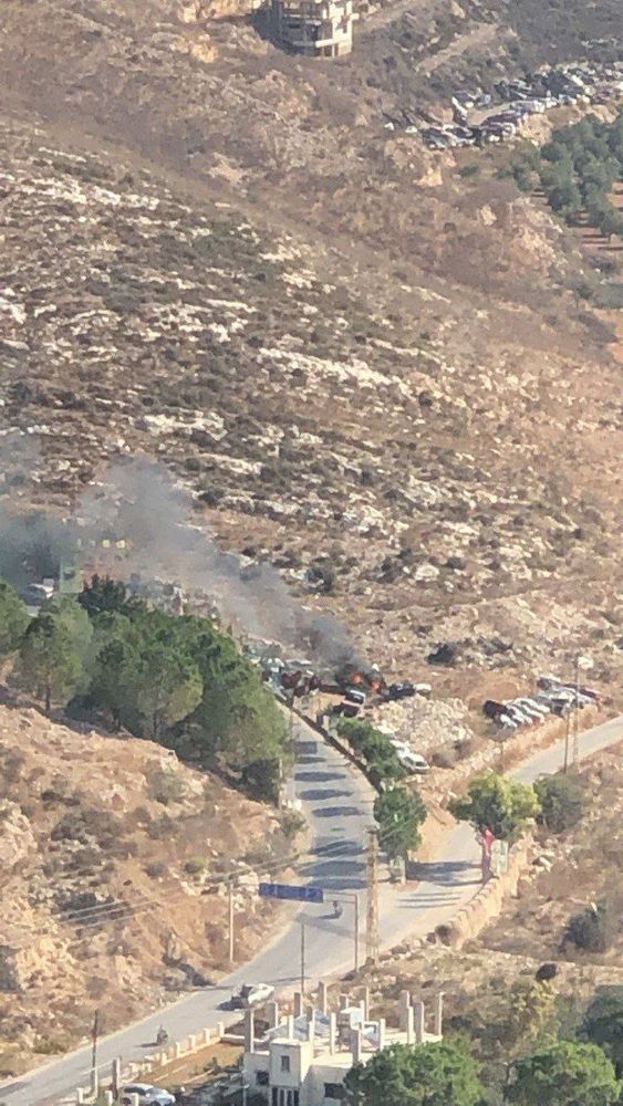 A zoomed-out aerial view of the same winding road. A plume of gray smoke rises from a burning vehicle near the road’s edge. Multiple cars are parked along the roadside and on the surrounding rocky terrain. The landscape is rugged, with dry hills and sparse trees.