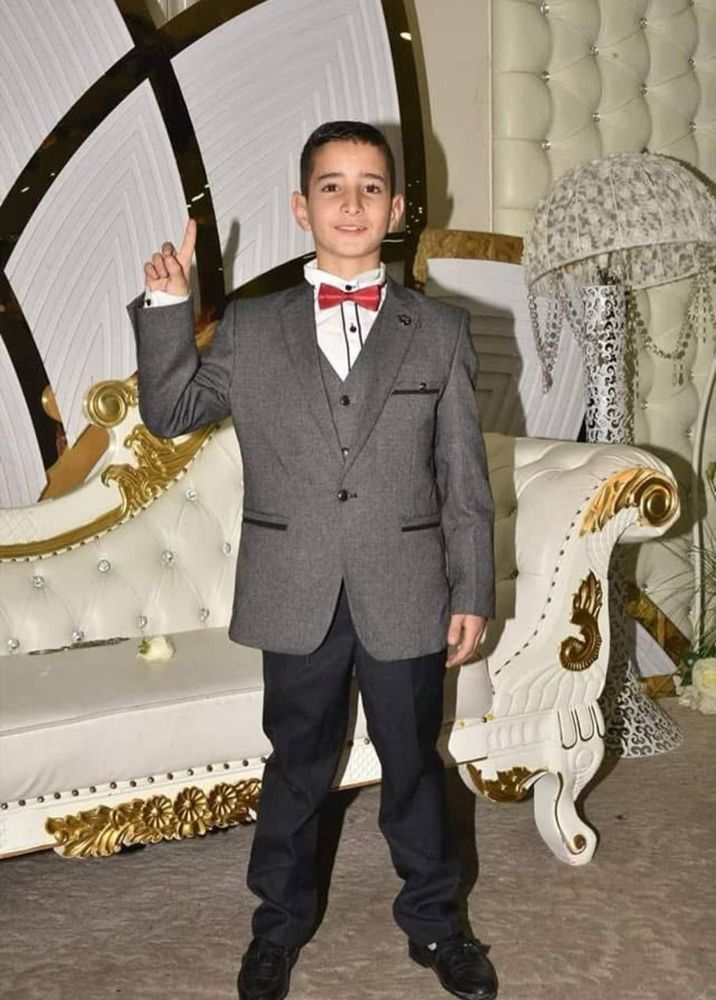 This photo shows a young boy dressed formally in a grey suit with a red bow tie, standing indoors and raising one finger while smiling softly at the camera. The background includes ornate white and gold furniture, suggesting a celebratory or formal occasion.