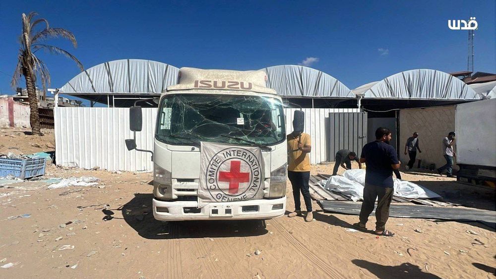 The image shows a white Isuzu truck bearing the emblem of the International Committee of the Red Cross (ICRC) on its front, with a cracked windshield and traces of dust. Several men stand around it — one holding a phone, others handling what appear to be white body bags laid on the sandy ground beside the vehicle. Behind them are metallic structures and white tarp-covered shelters under a clear blue sky.