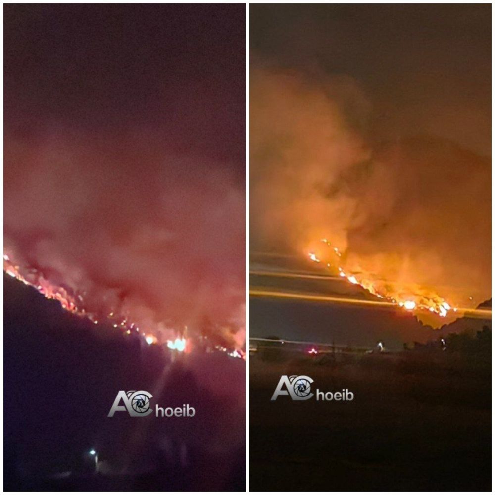 Two side-by-side nighttime photos show a mountainside engulfed in flames. In both images, long lines of fire stretch across the slope, creating a bright orange glow that lights up thick smoke rising into the dark sky. The fire appears intense and widespread, with multiple points of ignition visible along the ridge. In the foreground, faint silhouettes of buildings or structures can be seen near the bottom edge, indicating that residential areas are close by. The images are watermarked with the name “AChoieb.”