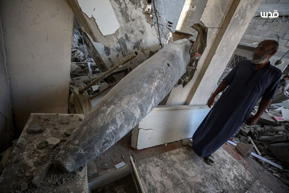 A closer angle shows the massive bomb embedded in a wall amid rubble and shattered tiles, with the same man standing nearby, his expression solemn.