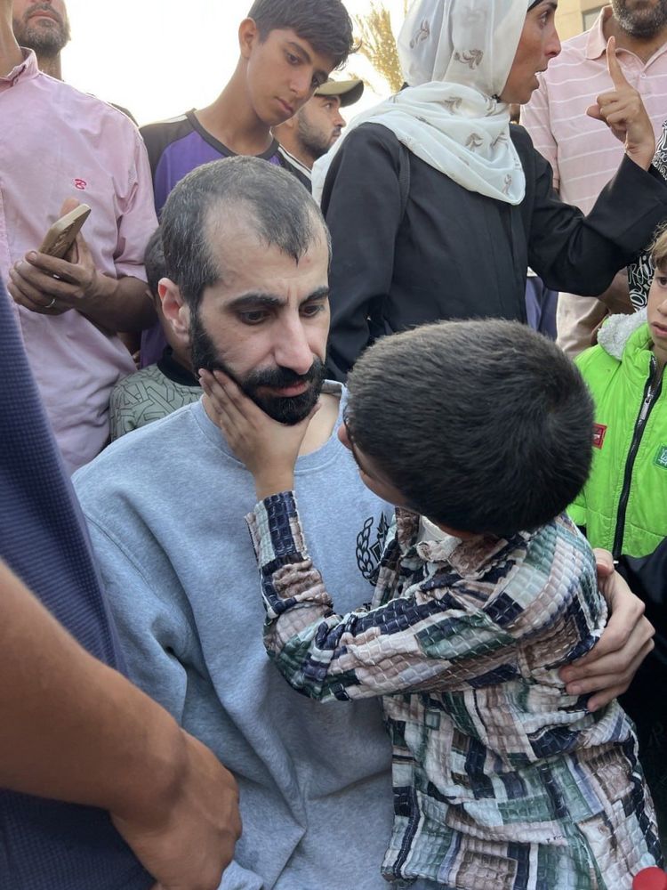 The image shows a deeply emotional reunion. A Palestinian man—identified as journalist Ahmad Abdul Aal—sits surrounded by a crowd, wearing a grey sweatshirt and looking somber, visibly affected. In the foreground, a young boy gently cups his face with both hands, gazing up at him with tenderness and concern. Around them, several people look on, some with solemn or tearful expressions. The moment captures both the exhaustion and relief of Abdul Aal’s release after enduring months of Israeli imprisonment, and the quiet strength of familial love and resilience in the aftermath.