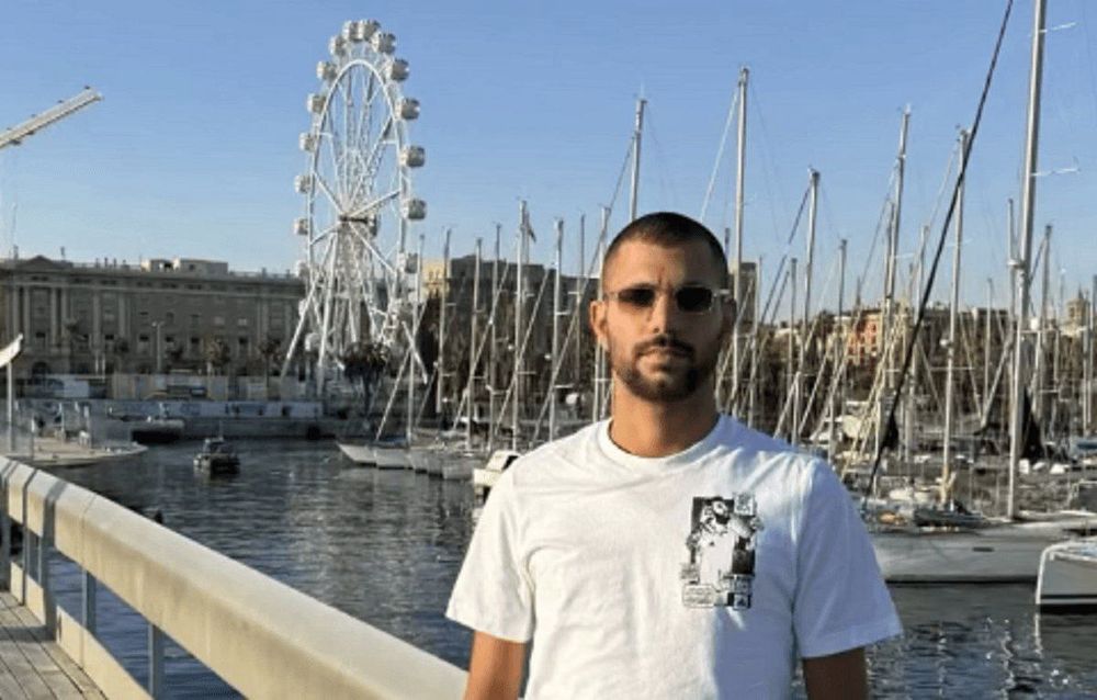 A man wearing sunglasses and a white T-shirt stands on a marina walkway. Behind him are sailboats, masts, and a large Ferris wheel against a clear sky. The scene appears calm and touristic.