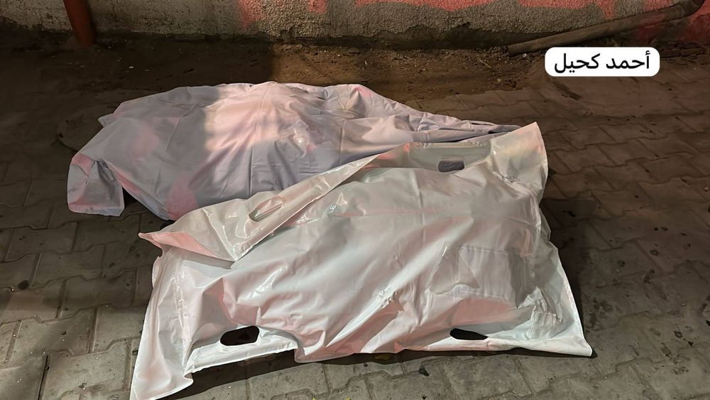 The image shows two white body bags laid side by side on a paved surface, illuminated by artificial light, possibly from emergency vehicles or nearby street lighting. The wall behind them is faintly marked with red stains or graffiti.