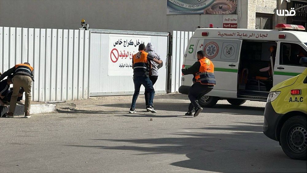 Paramedics wearing orange vests help a wounded man toward an ambulance outside a medical building, while another medic kneels beside a person on the ground.