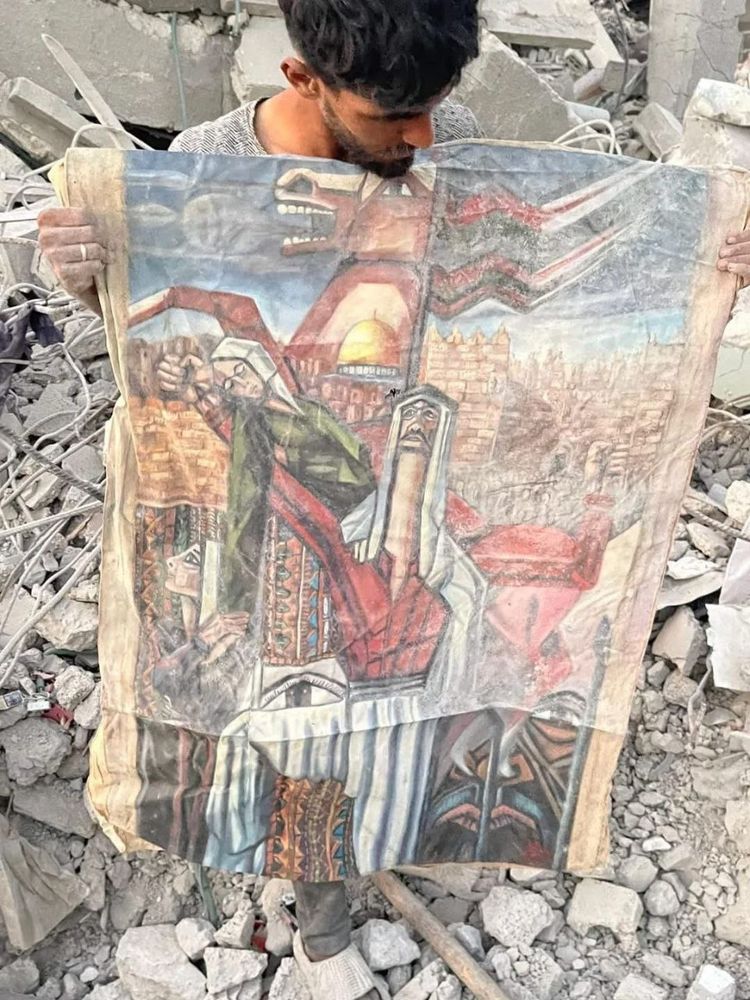 The image shows a man standing amid piles of rubble and shattered concrete, his hands dusty as he holds up a large, weathered painting. The artwork, titled “Identity” by late Palestinian artist Fathi Ghaben, depicts deeply symbolic figures in traditional Palestinian attire, with the Dome of the Rock visible in the background. Despite being creased and dirt-stained, the painting’s vivid reds, blues, and earth tones remain striking.

The man’s somber expression and the destruction surrounding him evoke a powerful sense of loss and resilience — the recovery of his father’s painting from the ruins a year after his father’s death underscores both personal and cultural survival amid devastation