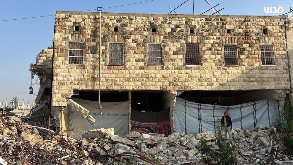 The front of a two-story stone building, part of the historic school, stands with its lower level blown open. Large sheets of tarp cover the exposed interior. Rubble is piled high around the base: broken stone blocks, twisted metal, shattered concrete. A person stands amid the destruction, underscoring the scale of the ruins.