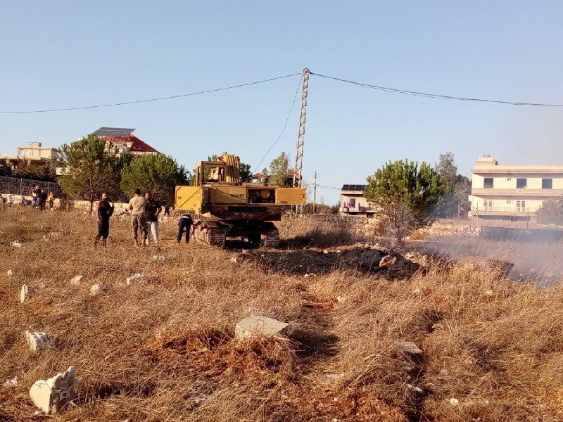 A yellow bulldozer sits in a dry, grassy field surrounded by several people inspecting the area. Some appear to be examining damage near the machine. Thin smoke drifts across the right side of the image, and scattered debris is visible on the ground. In the background are trees, power lines, and several residential houses under clear daylight. The scene has a tense, aftermath-like atmosphere.
