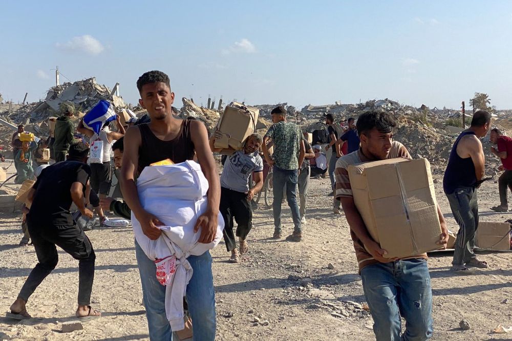 A group of Palestinian men walk through a dusty area filled with rubble and debris from destroyed buildings. They are carrying boxes, bags, and bundles of aid supplies in their arms, some struggling under the weight. The ruins of bombed structures rise in the background under a clear blue sky, showing the aftermath of heavy destruction. The expressions on their faces reflect exhaustion and urgency as they move through the devastated landscape.