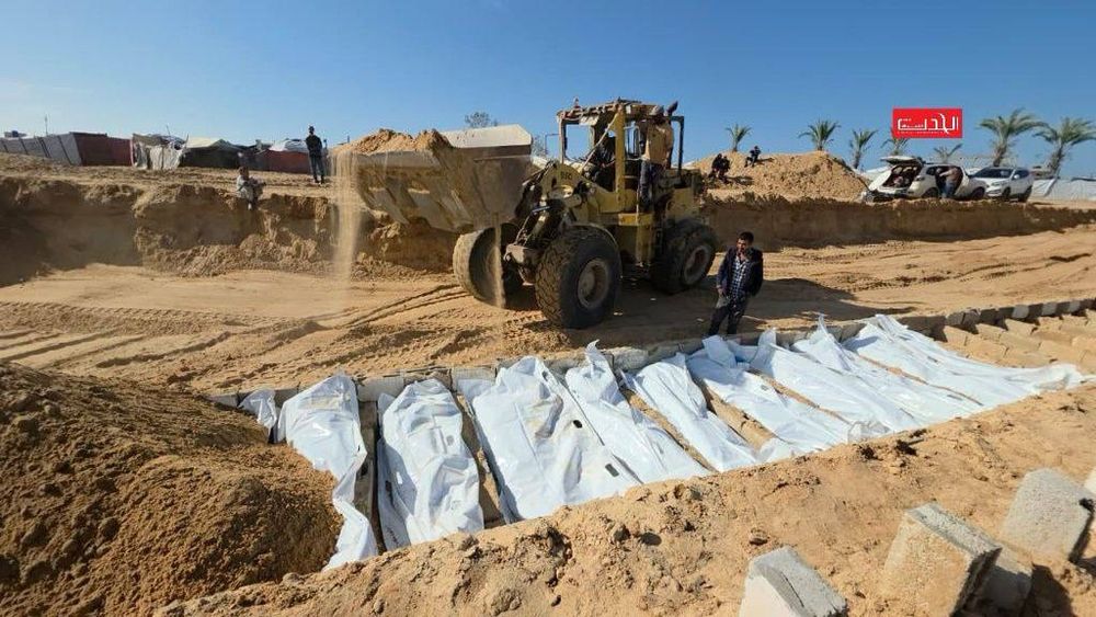 A mass burial site in Deir al-Balah shows rows of white body bags laid side by side inside a long trench dug into sandy ground. A bulldozer pours sand over the bodies as men stand nearby, watching or assisting. The area is surrounded by makeshift tents and displaced families’ shelters.