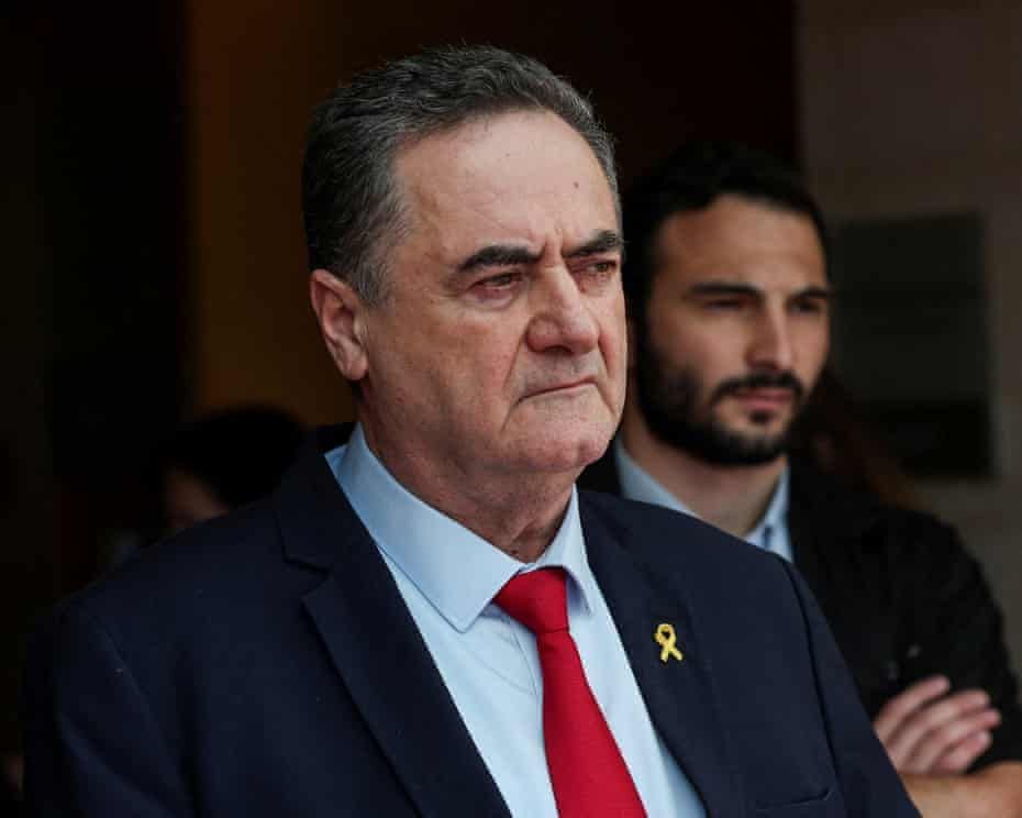 A man in a dark navy suit and red tie stands with a stern, serious expression, looking slightly to the side. A small yellow ribbon is pinned to his lapel. Behind him, another man in a dark jacket stands out of focus, his gaze directed forward. The lighting is natural, casting shadows that emphasize the main figure’s furrowed brow and resolute demeanor.