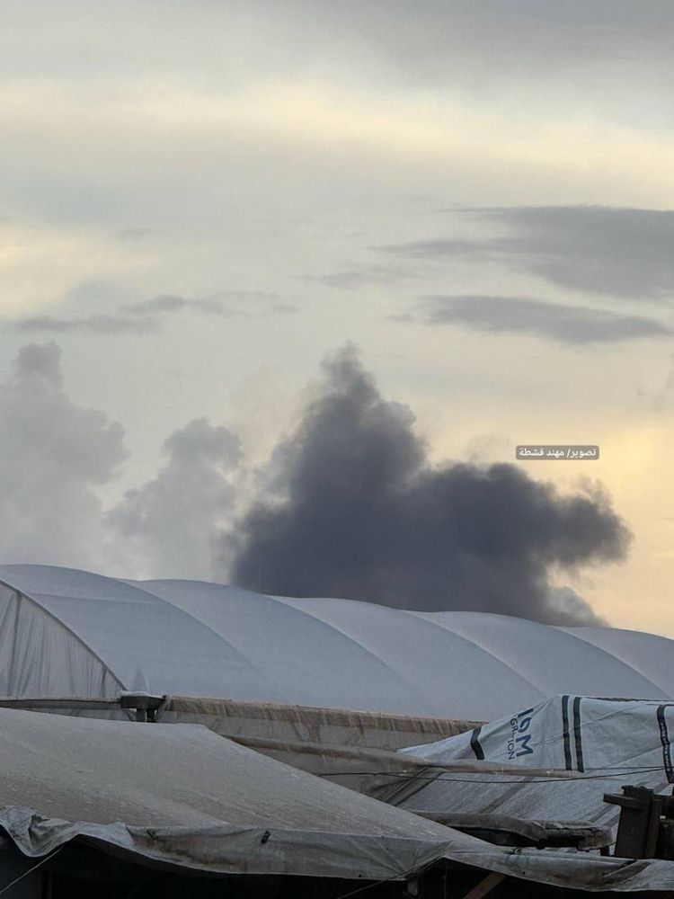 A thick plume of dark grey smoke rises into the cloudy evening sky in the distance. In the foreground are rows of large white tent-like shelters and tarpaulin-covered structures, suggesting a displaced persons camp. The smoke appears to come from an airstrike northwest of Rafah in southern Gaza.