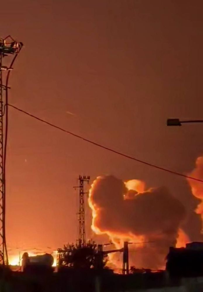  A massive nighttime explosion engulfs the sky in flames and thick orange smoke. Power lines and transmission towers are silhouetted against the intense fire, suggesting the blast occurred near an industrial or power facility.