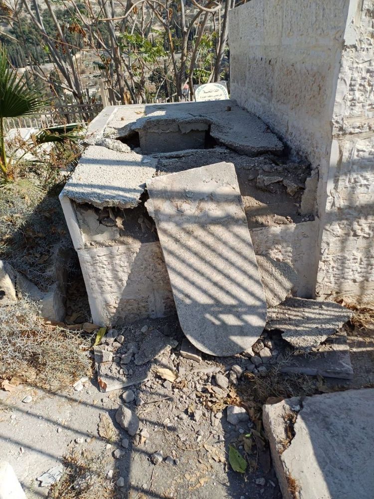 A close-up photo shows a damaged grave structure made of stone and concrete. The top slab has been broken into large pieces, leaving a jagged opening exposing the inside of the tomb. Broken chunks of concrete are scattered on the ground around it, along with dry leaves and debris. A metal railing casts shadows across the damaged grave, and trees and other graves are visible in the background.