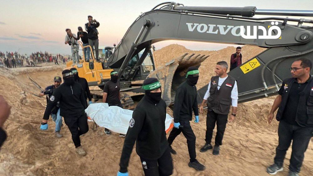 Masked men carry a white body bag near an excavator on sandy ground in Gaza, as others look on and a crowd gathers in the background during the recovery of bodies from beneath the rubble.
