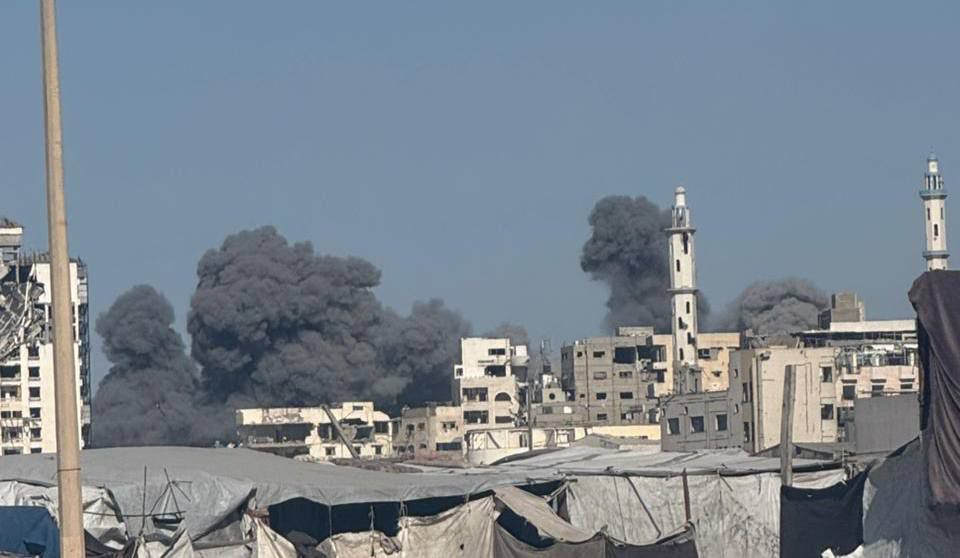 Thick black smoke rises in towering plumes from multiple explosions across Gaza City. Apartment blocks and minarets of mosques stand in the foreground, while makeshift tents are visible at ground level, underscoring the devastation hitting the Al Shati refugee camp.