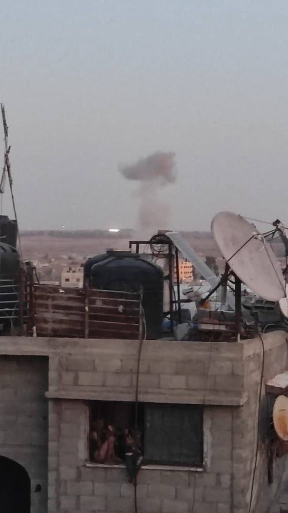 A plume of grey smoke rises in the distance over the eastern area of Al-Bureij Camp in central Gaza. In the foreground, the rooftop of a concrete building is visible, cluttered with water tanks, satellite dishes, and cables. Below, several children can be seen looking out of a window, watching the explosion from inside the building. The sky is hazy, and the scale of destruction is visible on the horizon.