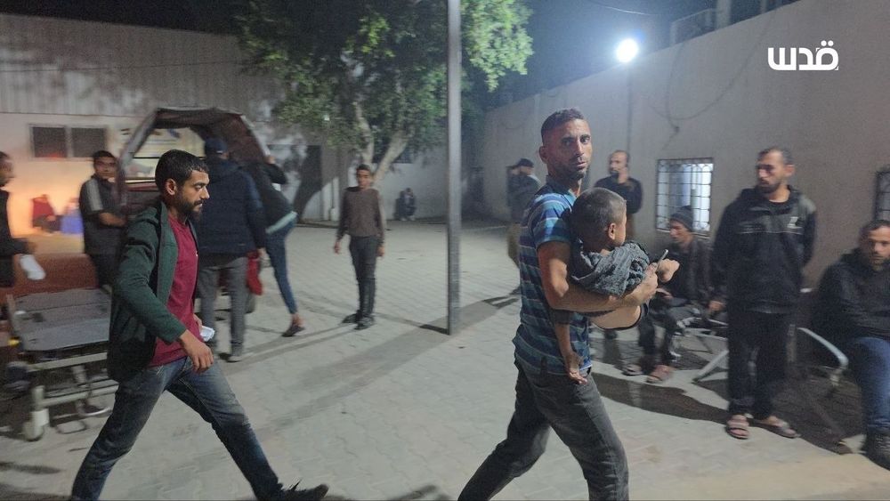 A man in a striped shirt carries a young child in his arms while moving quickly across a courtyard at night, as several people stand or walk around in the background under harsh emergency lighting.