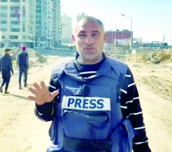 A journalist stands outdoors wearing a blue protective vest labeled “PRESS” on the front. He faces the camera with one hand slightly raised as if speaking or gesturing. Behind him are damaged streets, scattered debris, and tall residential buildings, suggesting he is reporting from a conflict-affected area. A few people walk in the background.