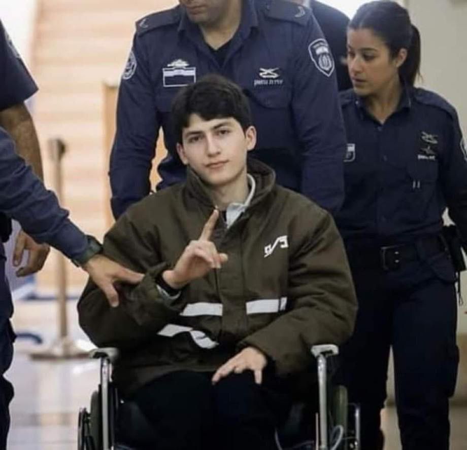 Two photos show a young Palestinian man in a wheelchair, escorted by multiple Israeli prison officers in dark blue uniforms. In the first image, he wears a brown jacket with white stripes and raises his right index finger calmly while being pushed through a corridor. 